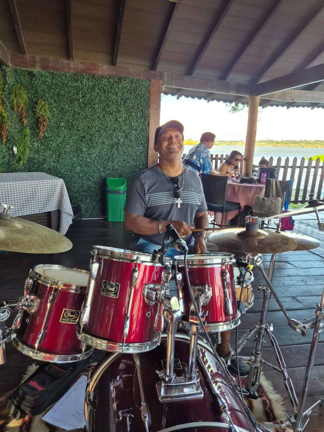 Baterista de boné sorrindo atrás da bateria vermelha, num deque coberto.