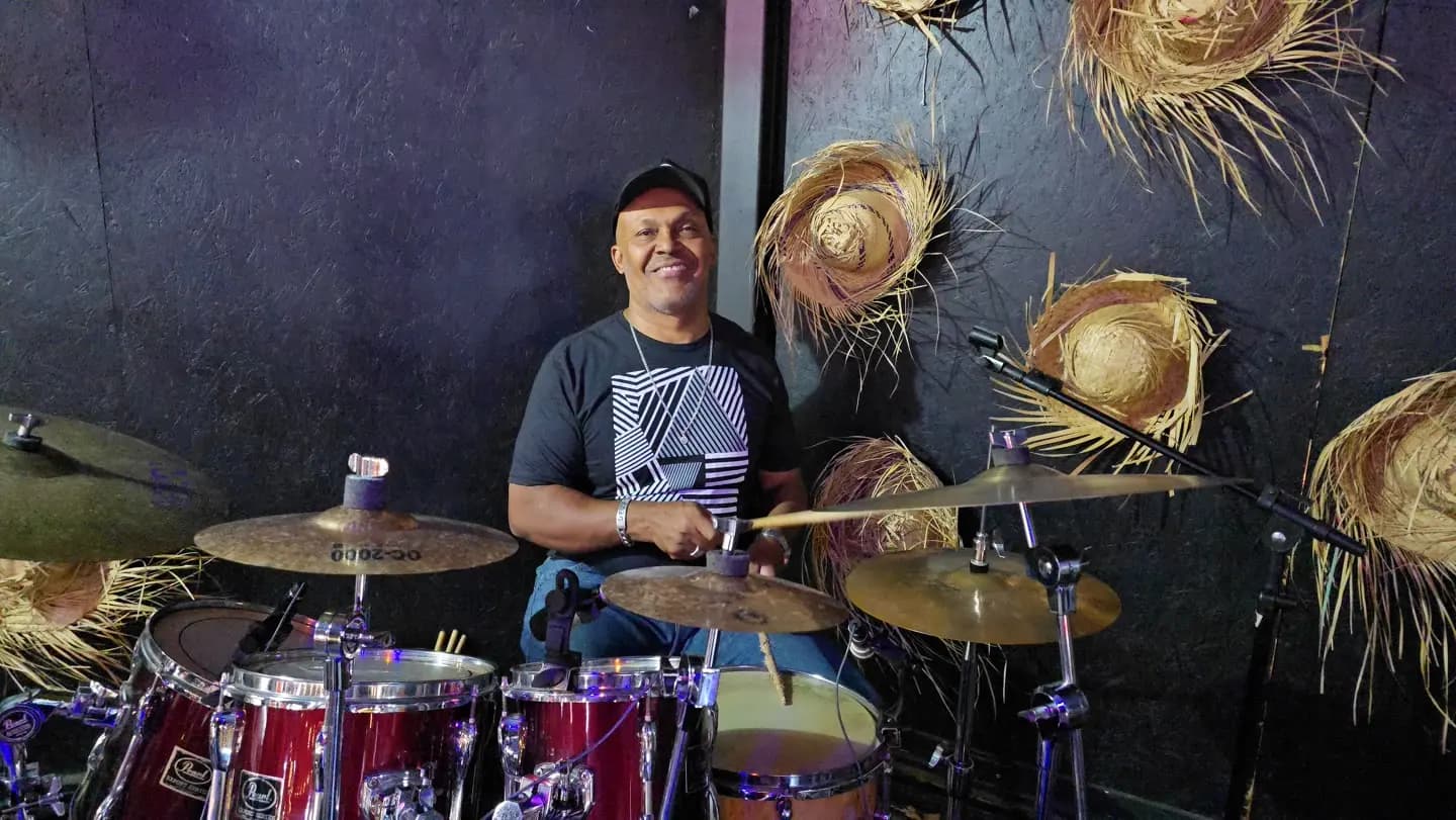 Baterista sorrindo atrás de uma bateria vermelha, com chapéus de palha na parede.