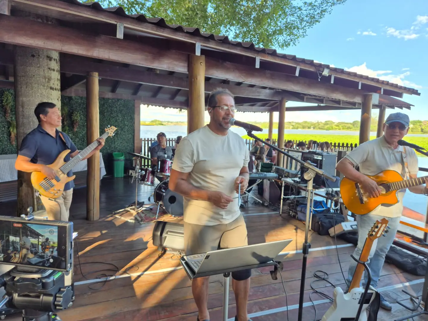 Baixista, cantor e violonista num deque de madeira, de dia, em festa de aniversário à beira do lago.
