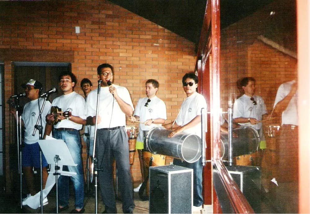 Foto antiga: a banda de camiseta branca num palco de tijolo aparente, com violão, voz, surdo e conga.