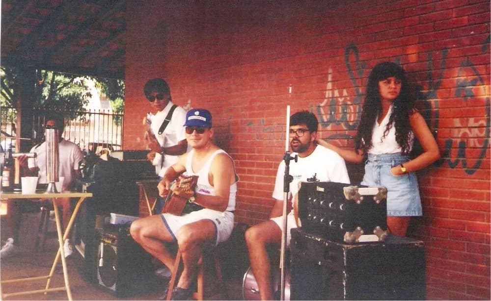 Foto antiga: quatro músicos e uma moça sob um telhado, diante de uma parede de tijolo grafitada, com violão e baixo.