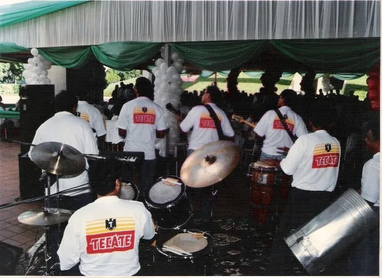 Foto antiga: a banda de costas, de camiseta branca da Tecate, tocando sob uma tenda com balões.